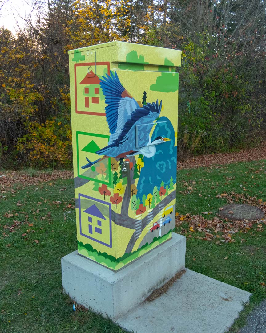 A mural on a traffic light box depicting a blue heron in flight over a forested neighbourhood.
